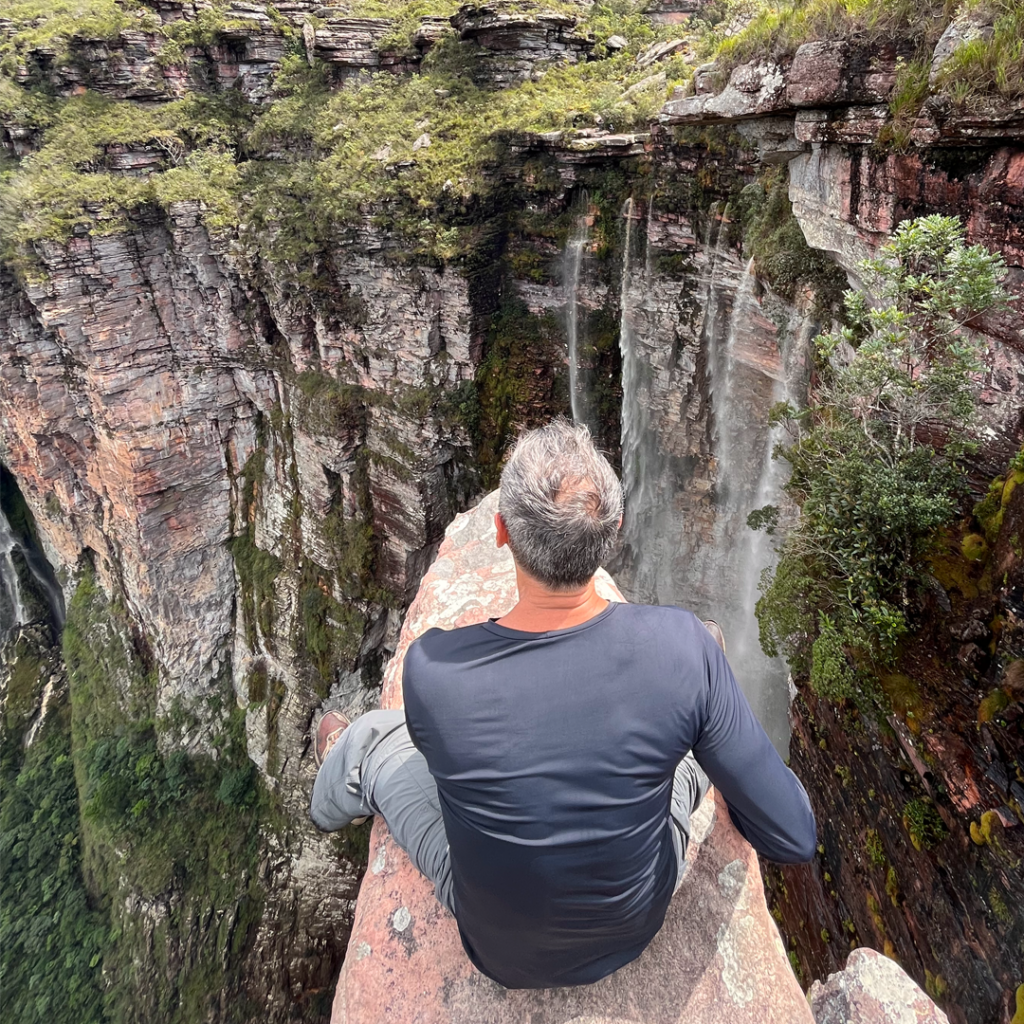 Homem sentado no Mirante do Cachoeirão no Vale do Pati, Chapada Diamantina. Cachoeira e vegetação no horizonte.