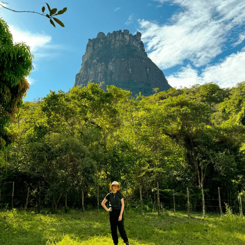 Mulher em roupa de trilha posa diante do Morro do Castelo, uma das principais atrações do Vale do Pati, Chapada Diamantina, rodeado por vegetação verde e céu azul.