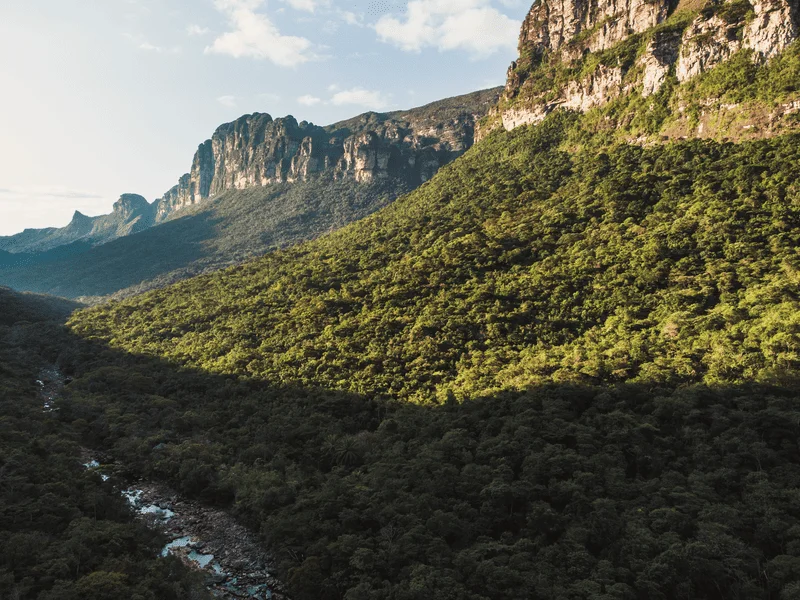 Drone sobrevoa área do rio Lapinha, na trilha do Vale do Pati, Montanhas laterais e vegetação com sol à pino ao fundo