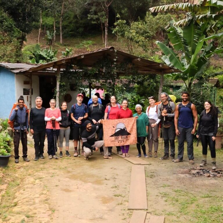 Grupo de turistas posando sorridentes lado a lado em frente a uma casa de apoio de paredes azuis e telhado rústico, segurando a bandeira laranja com o logo da Xandy Trekking no Vale do Pati.