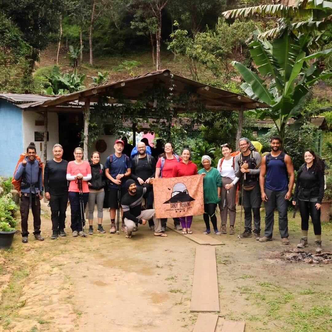 Grupo de turistas posando sorridentes lado a lado em frente a uma casa de apoio de paredes azuis e telhado rústico, segurando a bandeira laranja com o logo da Xandy Trekking no Vale do Pati.
