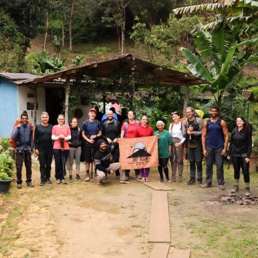 Descubra o Vale do Pati – Hospedagem Local | Xandy Trekking Grupo de turistas posando sorridentes lado a lado em frente a uma casa de apoio de paredes azuis e telhado rústico, segurando a bandeira laranja com o logo da Xandy Trekking no Vale do Pati.